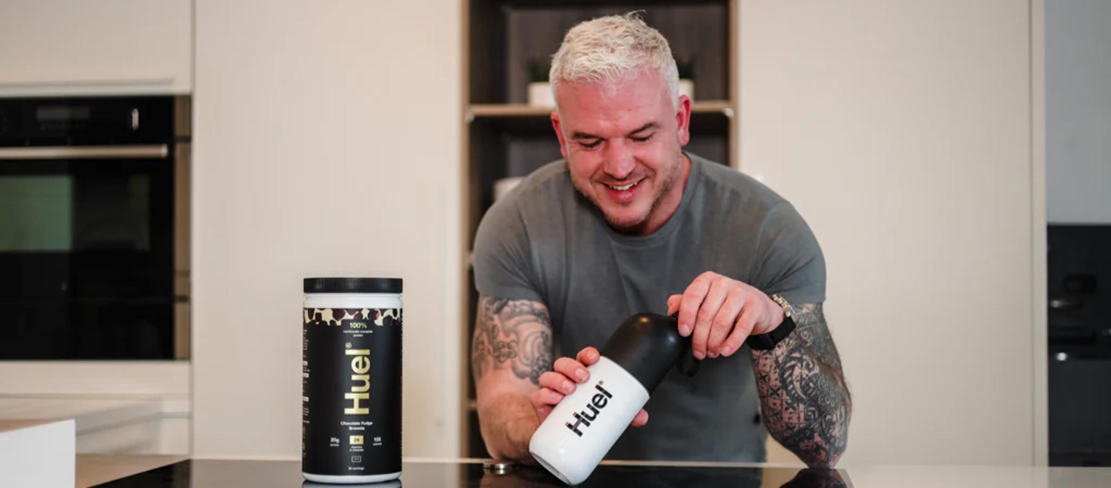 Man leaning on a kitchen worktop using Huel Complete Protein in a shaker
