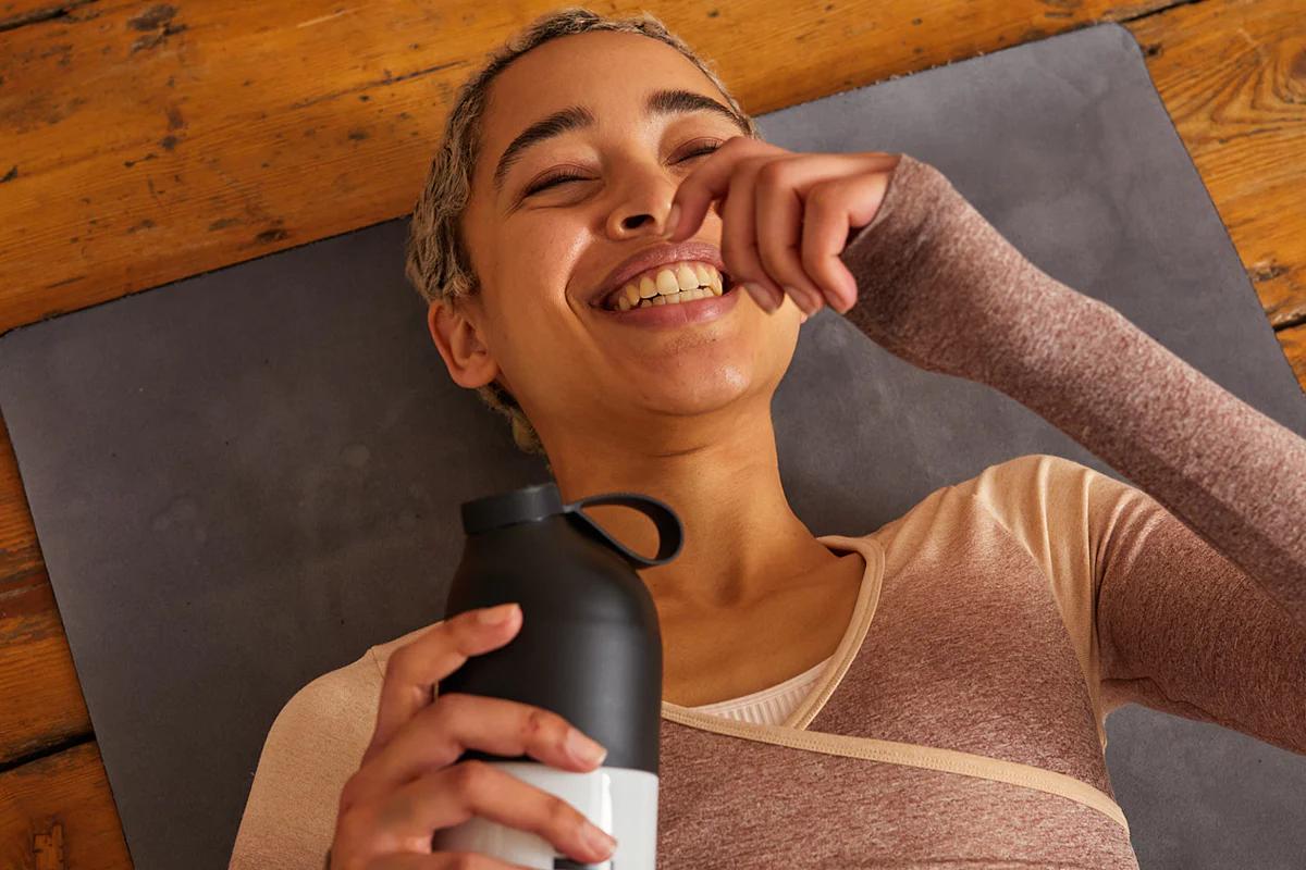 woman laying on a yoga mat laughing whilst holding a huel shaker