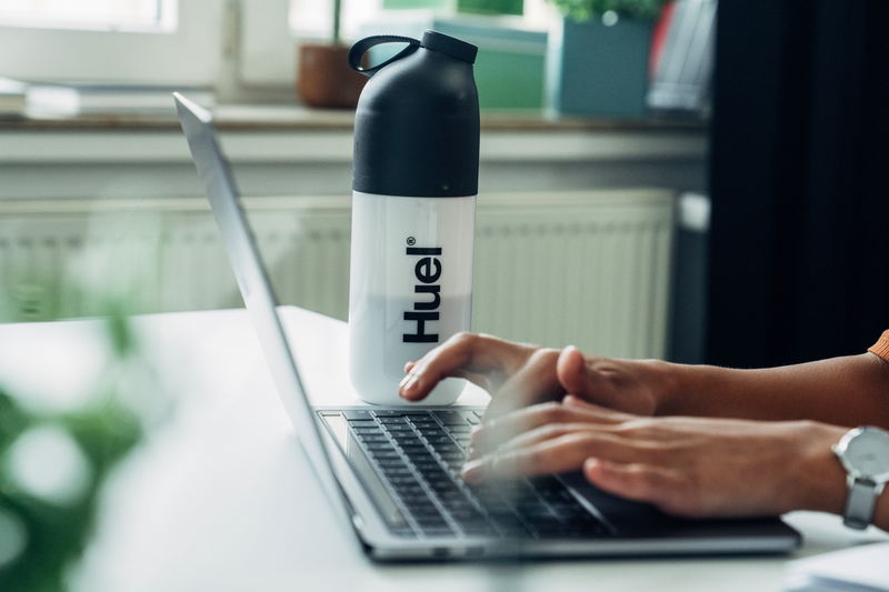 someone working on their laptop with a huel shaker on the desk besides them