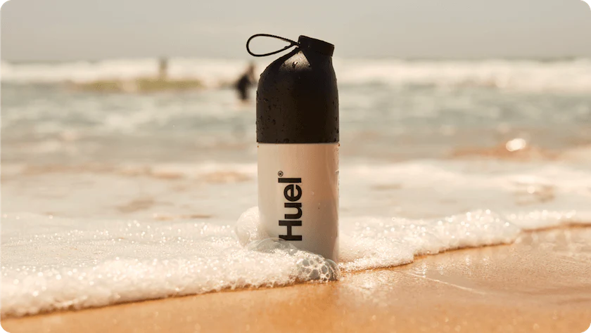 a huel shaker stood in the sand on a beach as a wave crashes around it