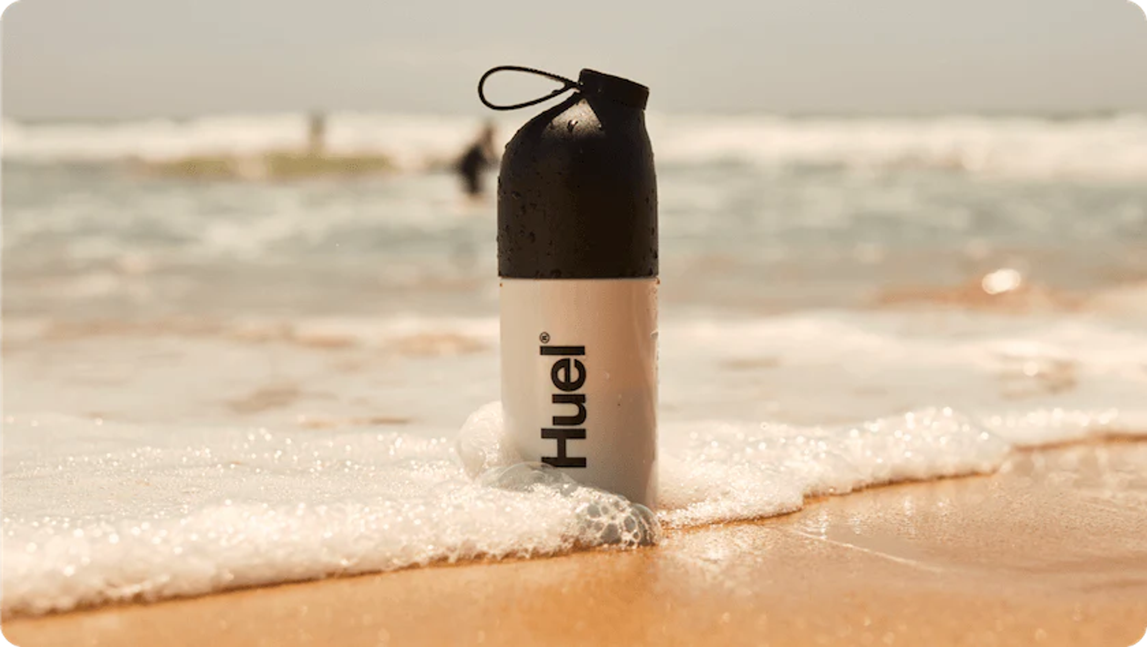 a huel shaker stood in the sand on a beach as a wave crashes around it