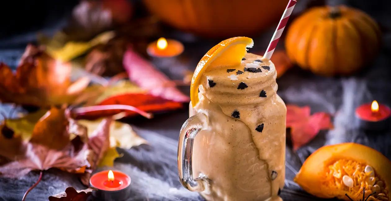 milkshake in a glass jar shaped mug with chocolate sprinkles a straw an orange wedge and pumpkins and autumn leaves in the background