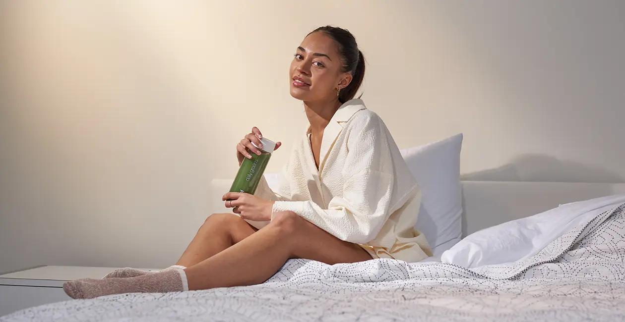 woman sat on bed holding Huel Daily Greens in a Huel bottle