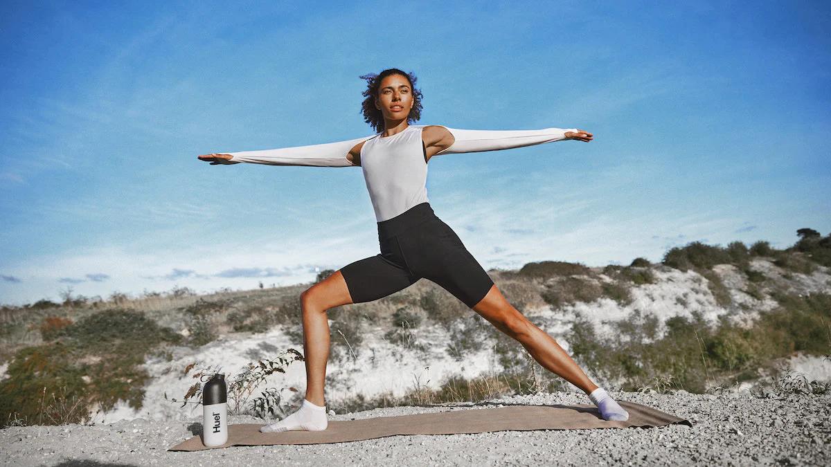 woman stretching and doing yoga outside with blue skies and fluffy clouds behind and a huel shaker on the ground next to her