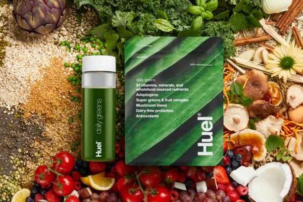 Huel Daily Greens Beutel und Flasche liegen auf einer bunten Auswahl verschiedener Obst- und Gemüsesorten