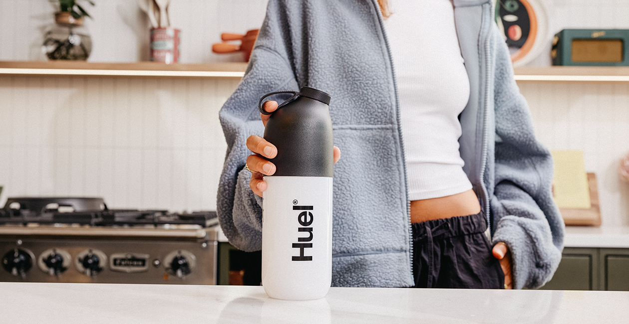 woman stood in a kitchen with her hand on a Huel shaker which is on the kitchen counter