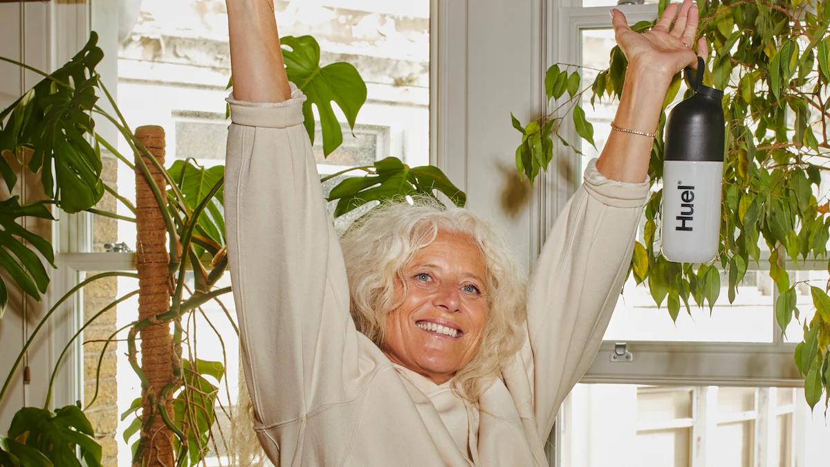 woman with her arms stretched up smiling holding a huel shaker next to a plant