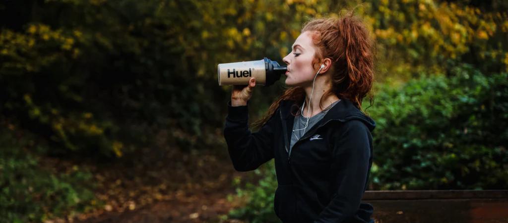 woman drinking from a huel shaker in the woods with earphones in
