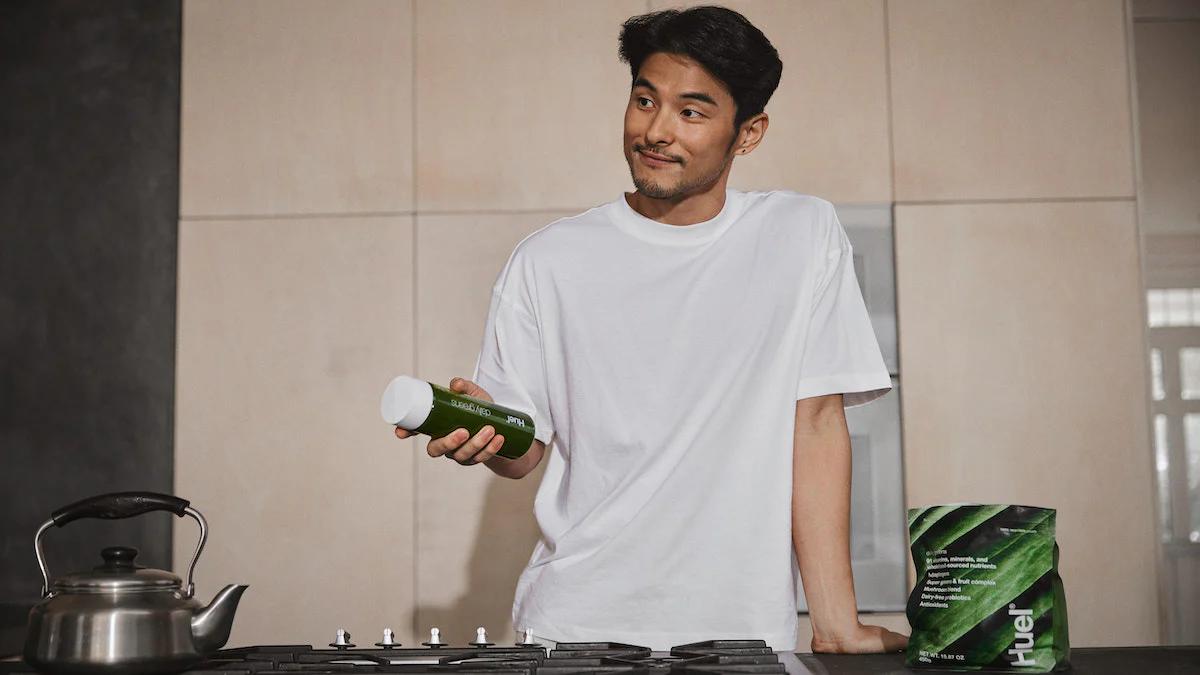 man stood in kitchen holding huel daily greens bottle with a pack of huel daily greens on the side