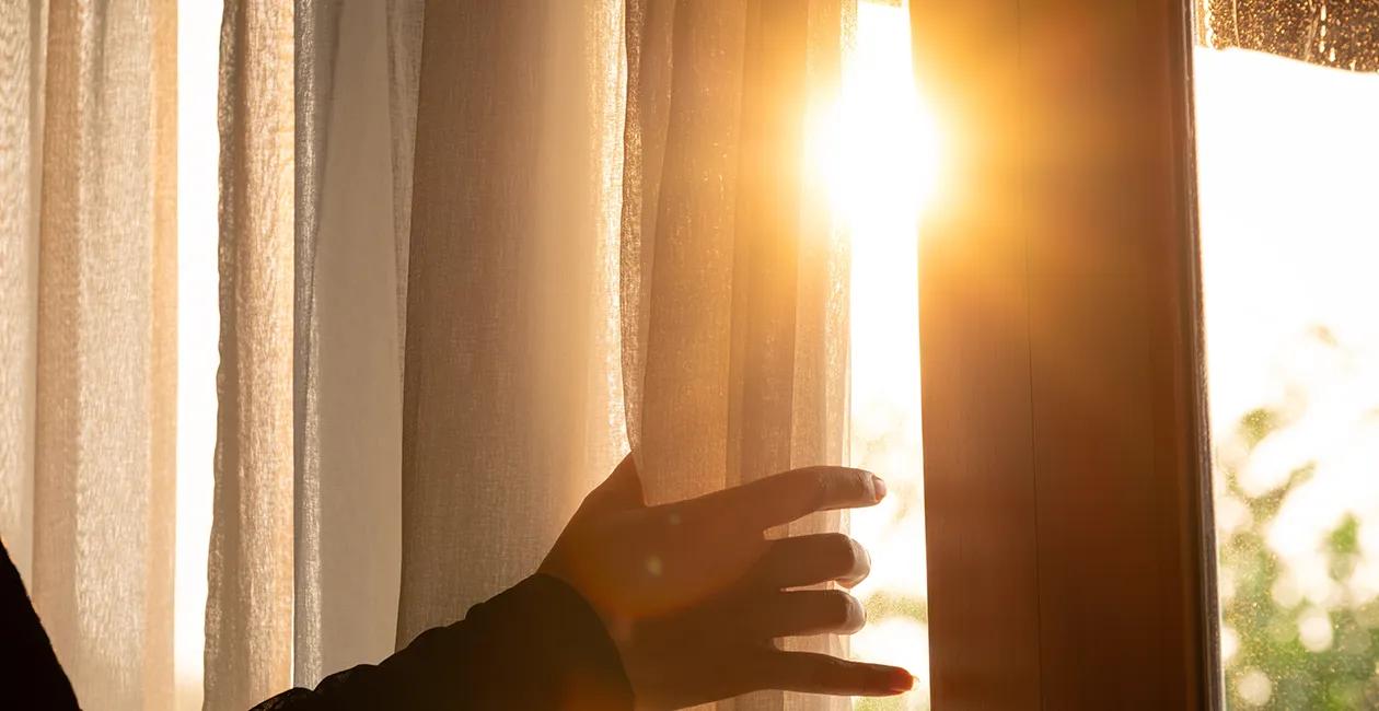 Sun shining through a window with a person holding the curtain