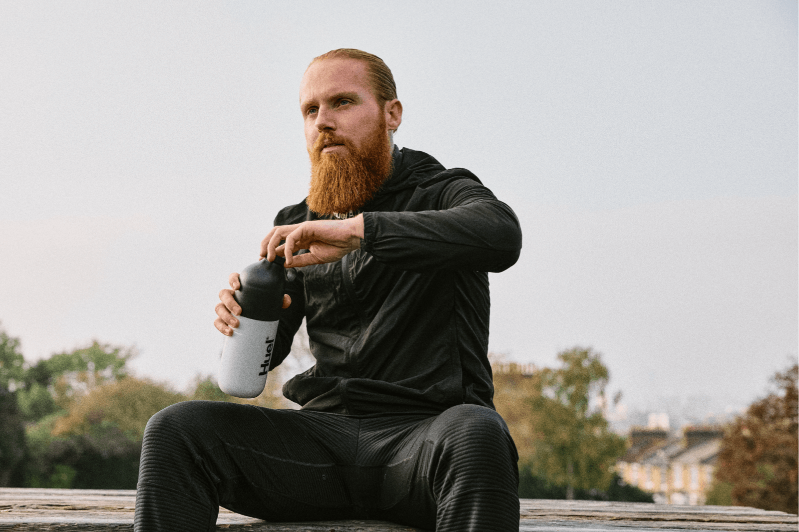 Russ Cook - The Hardest Geezer - sitting on a bench in a black tracksuit, opening a shaker of Huel