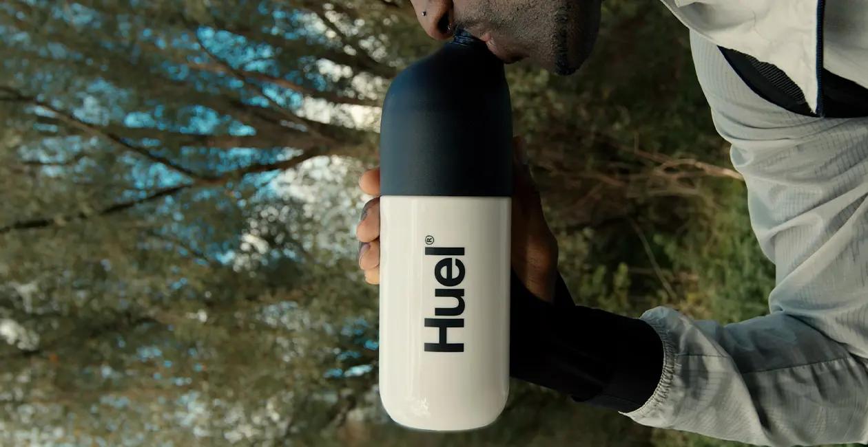 Huel shaker in the centre with a man holding and drinking from it while outside with trees in the background