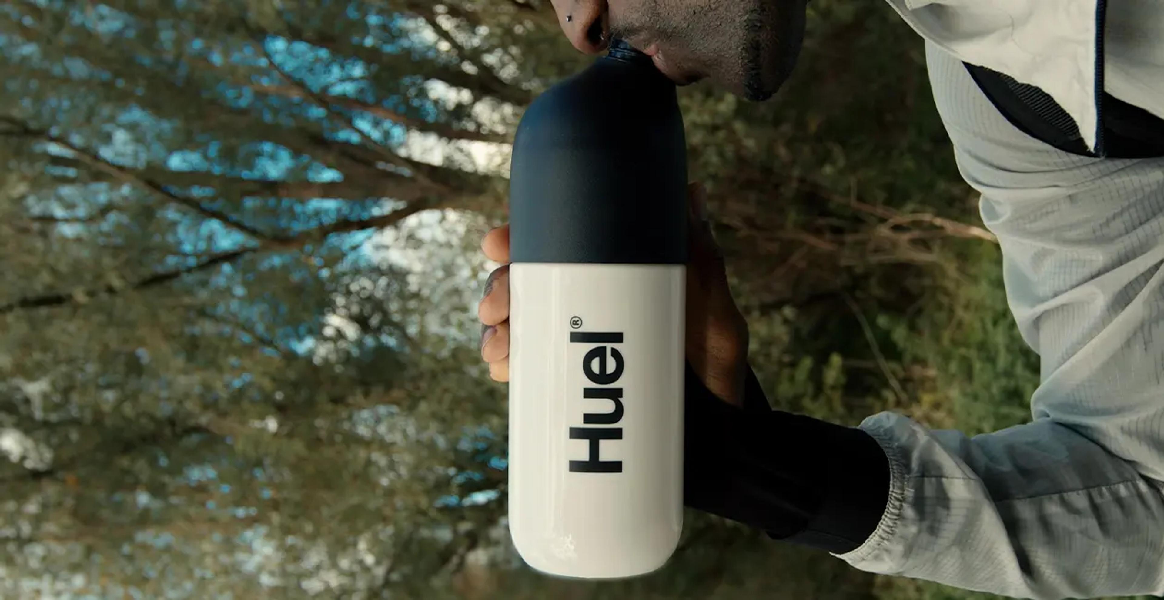 Huel shaker in the centre with a man holding and drinking from it while outside with trees in the background