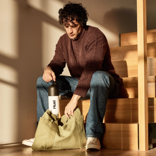 Un homme portant un pull en maille foncé et un jean est assis sur des escaliers en bois baignés de lumière, en train de ranger une bouteille Huel blanche et noire dans un grand sac en toile.