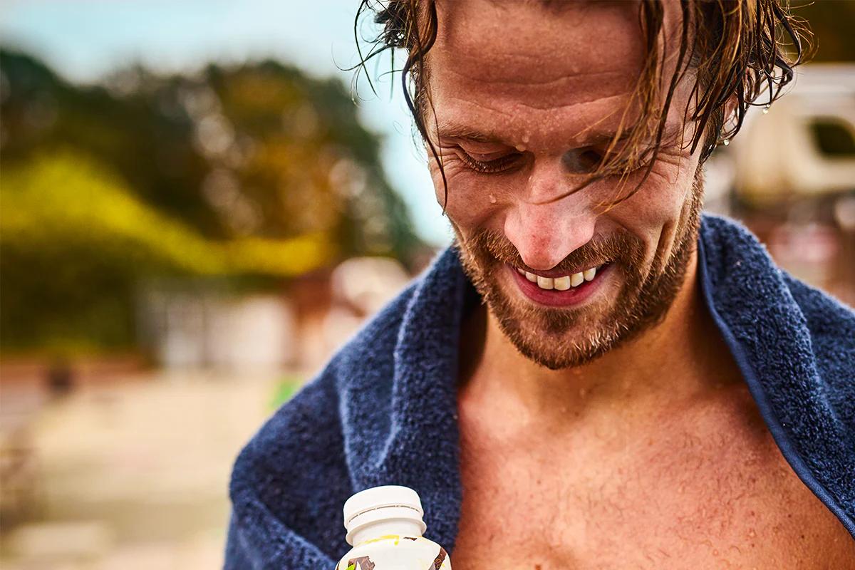 man outside who is wet and has a towel on his shoulders holding a huel ready-to-drink