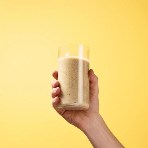 A hand holding a glass containing Huel Black Edition in Banana flavour on a yellow background