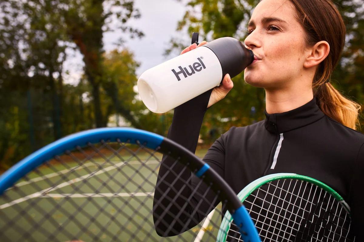 woman drinking from a huel shaker stood on a tennis court with a tennis racket under her arm