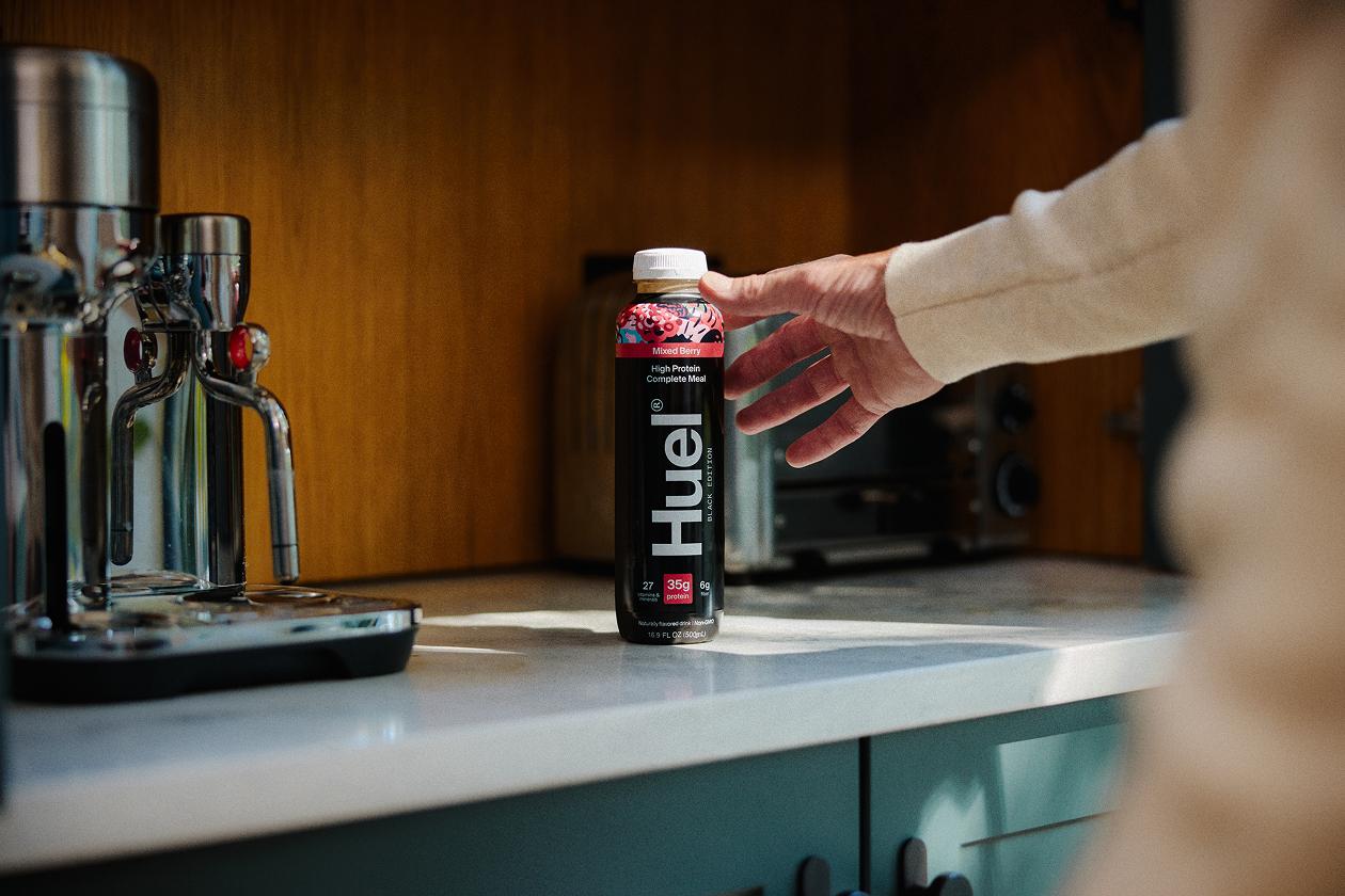 A Huel Black Edition Ready-to-drink in Mixed Berry flavor on a counter with a man reaching for it
