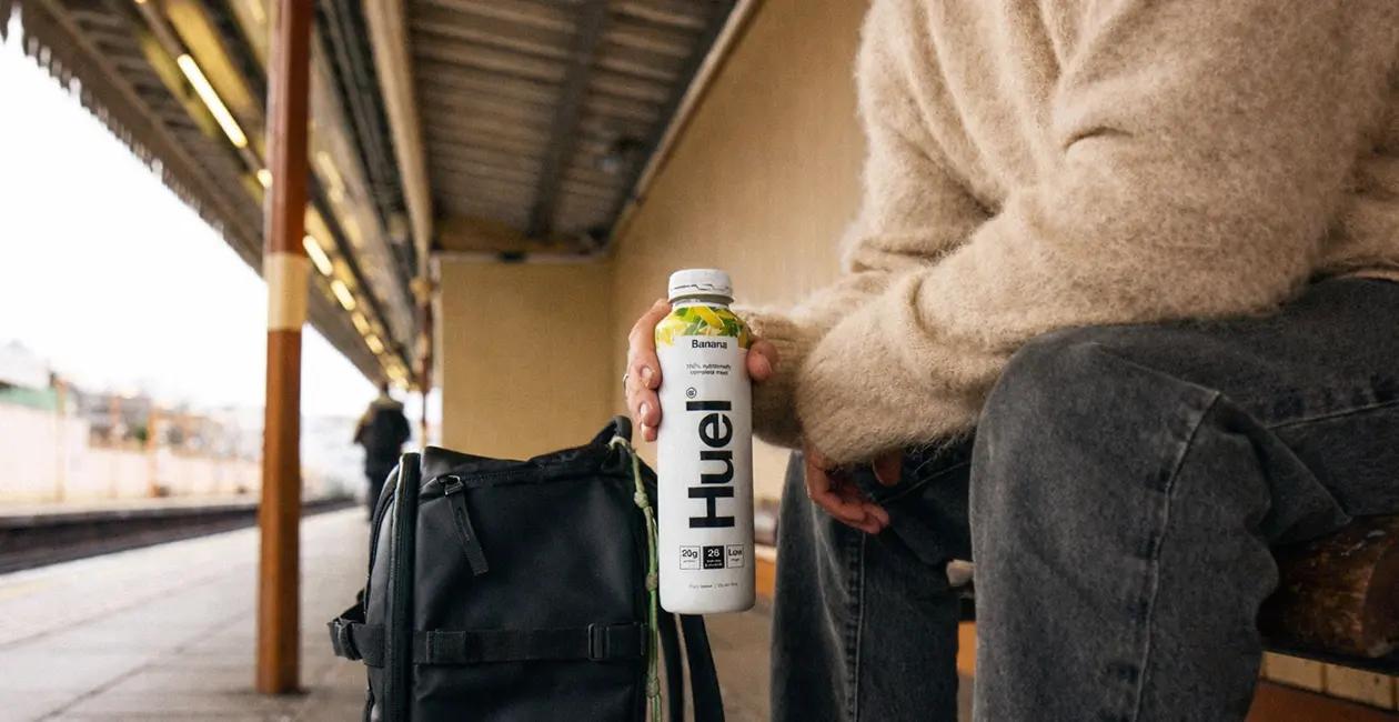 person sat on a bench at a train station holding a Huel Banana Ready-to-drink bottle with a bag next to them on the ground
