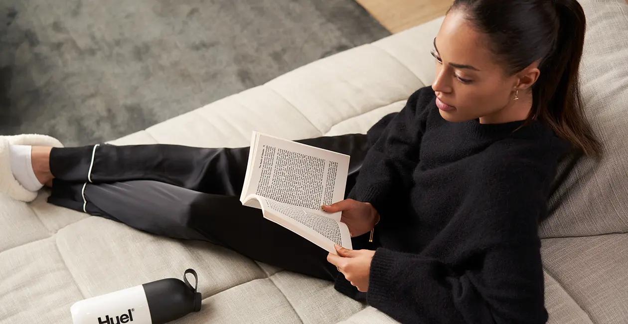 woman sat on sofa with her feet up reading a book with a Huel shaker on the sofa next to her