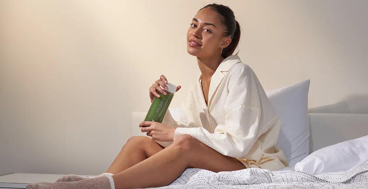 woman sat on a bed in an oversized shirt holding a Huel Daily Greens bottle looking at the camera