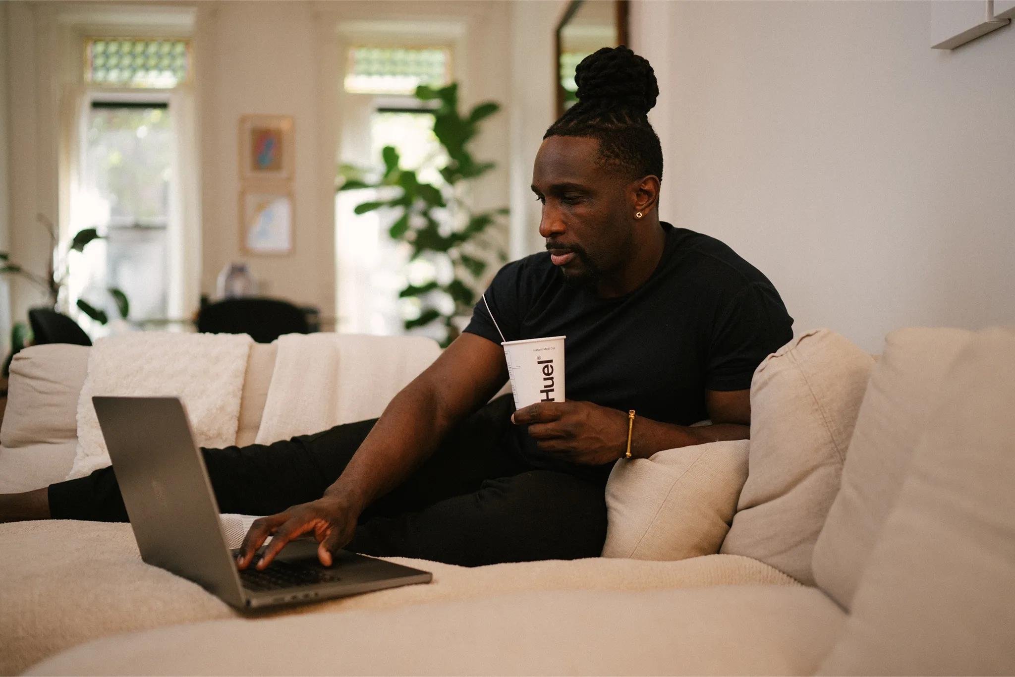 man sat on sofa eating a huel hot and savoury pot while working on his laptop