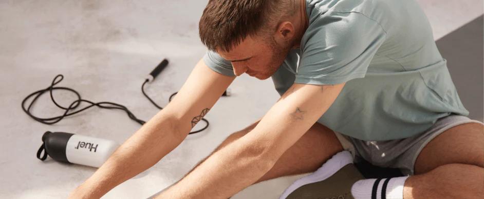 man sat on a yoga mat reaching down to his foot to stretch there is a skipping rope and huel shaker on the floor behind him