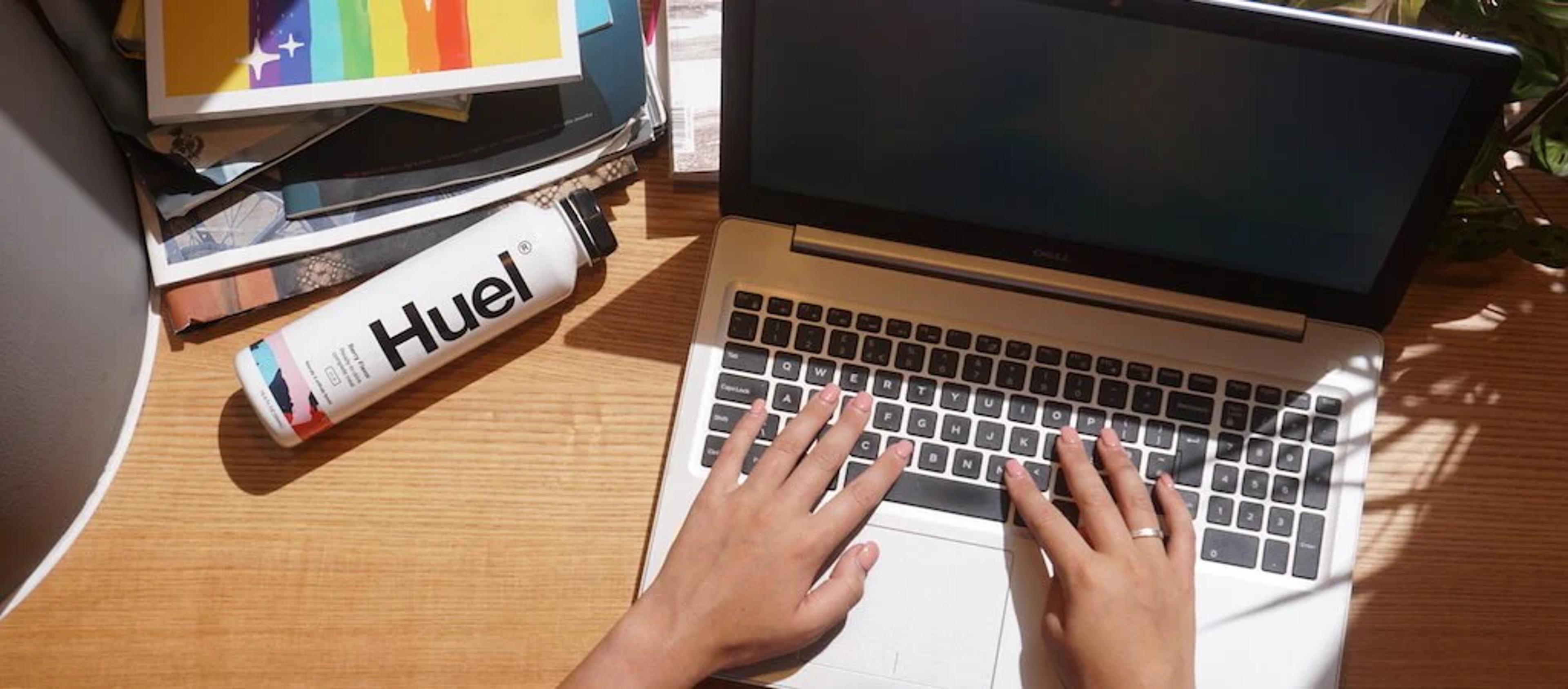 Huel Ready-to-drink on a desk next to someone typing on a laptop