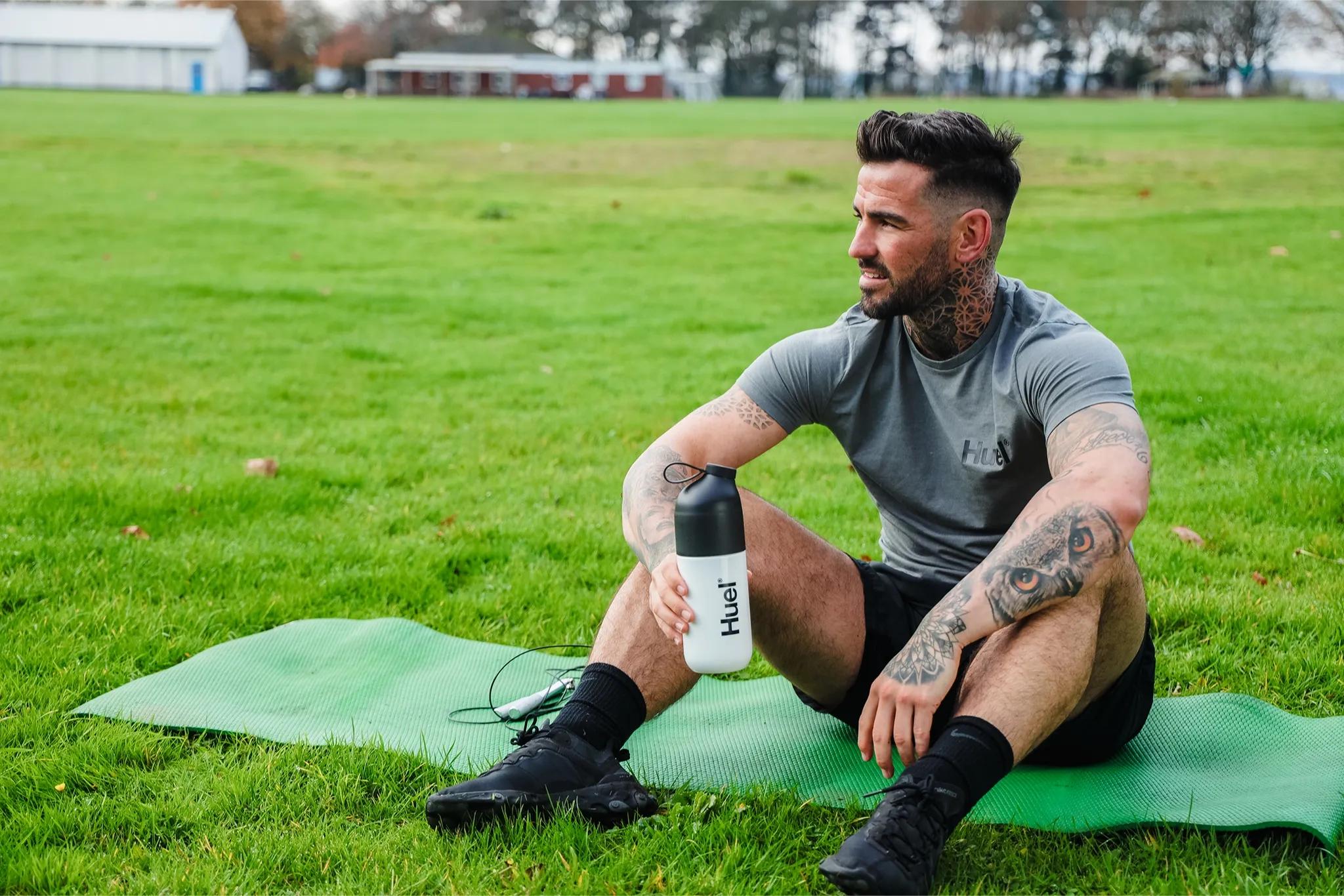 man sat on a yoga mat on the grass outside holding a huel shaker
