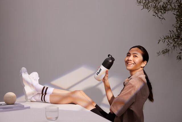 woman sat on a chair with her feet up on the table holding a huel shaker and smiling
