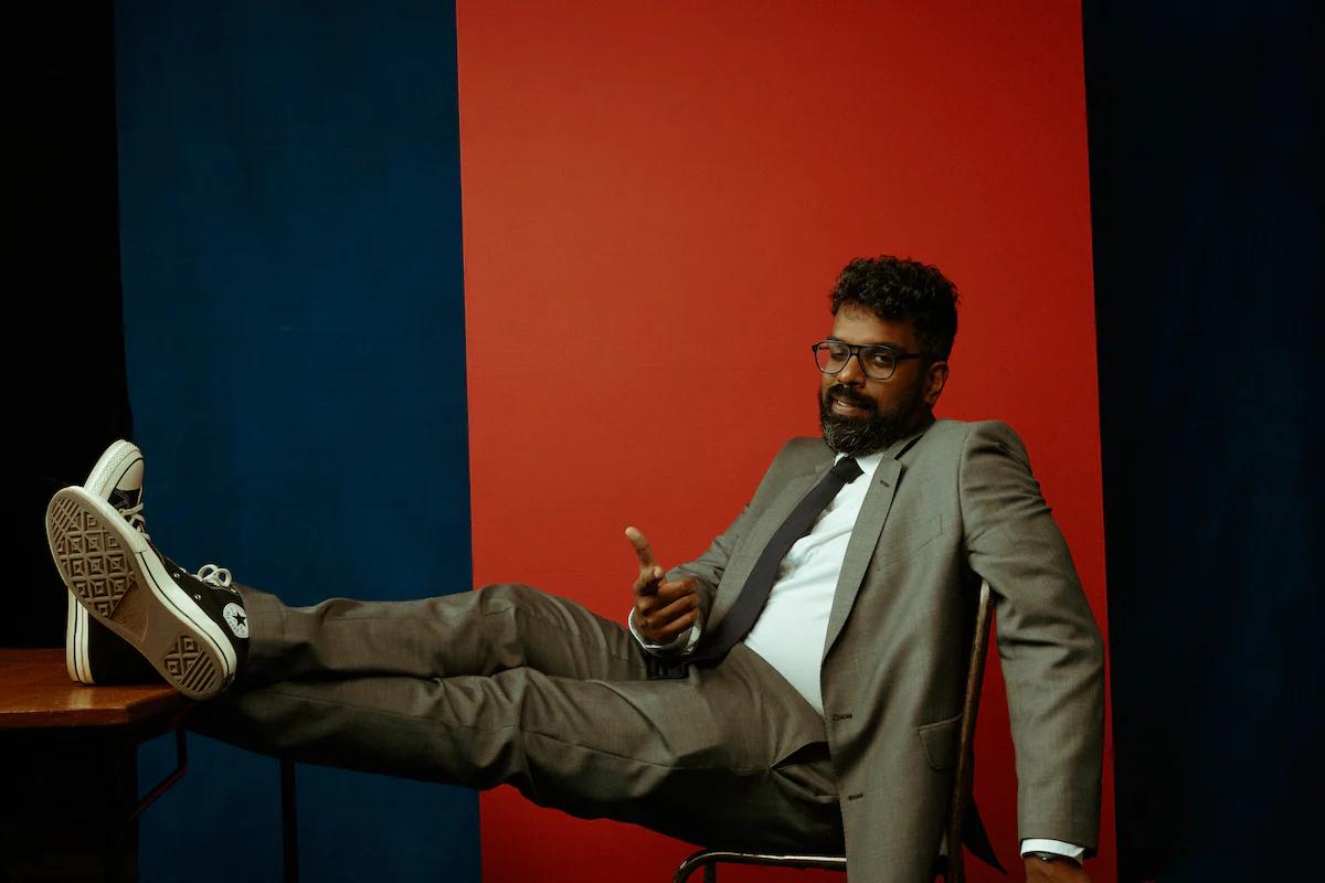 Romesh Ranganathan sat down with his feet up winking and pointing at the camera