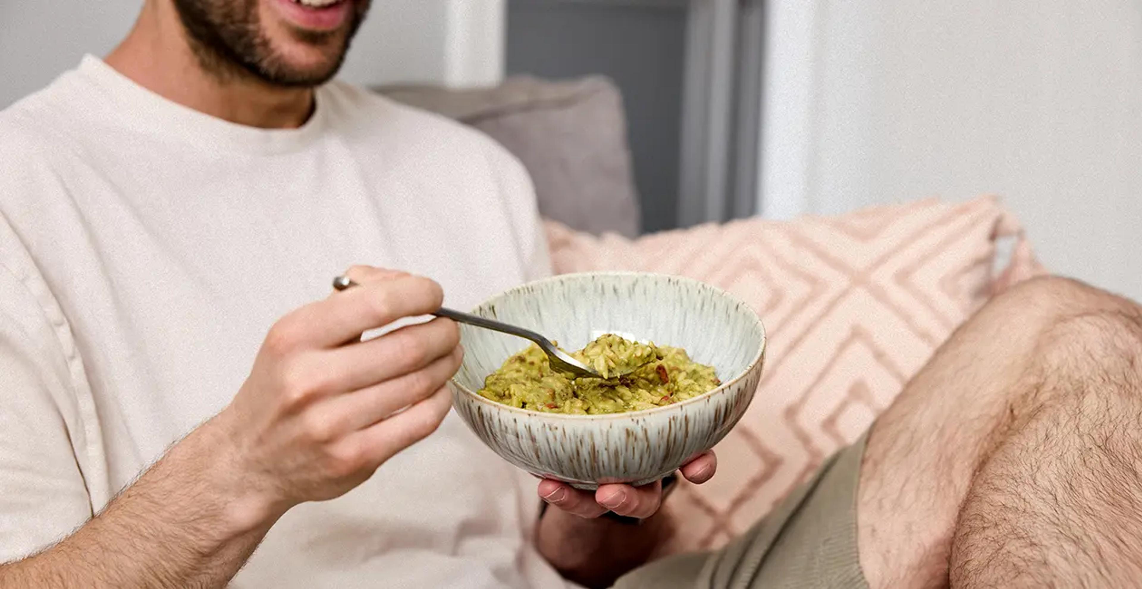 man sat on sofa eating huel hot and savoury from a bowl with a fork