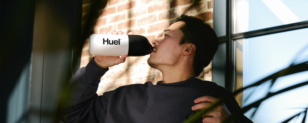 Image of a man reclining in a sunlit room on a chair while drinking from a black and white Huel shaker bottle