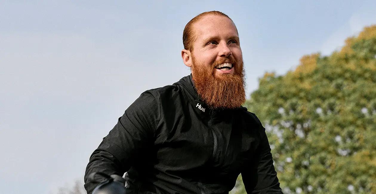 Russ Cook wearing a Huel jacket sat outside looking off into the distance and smiling