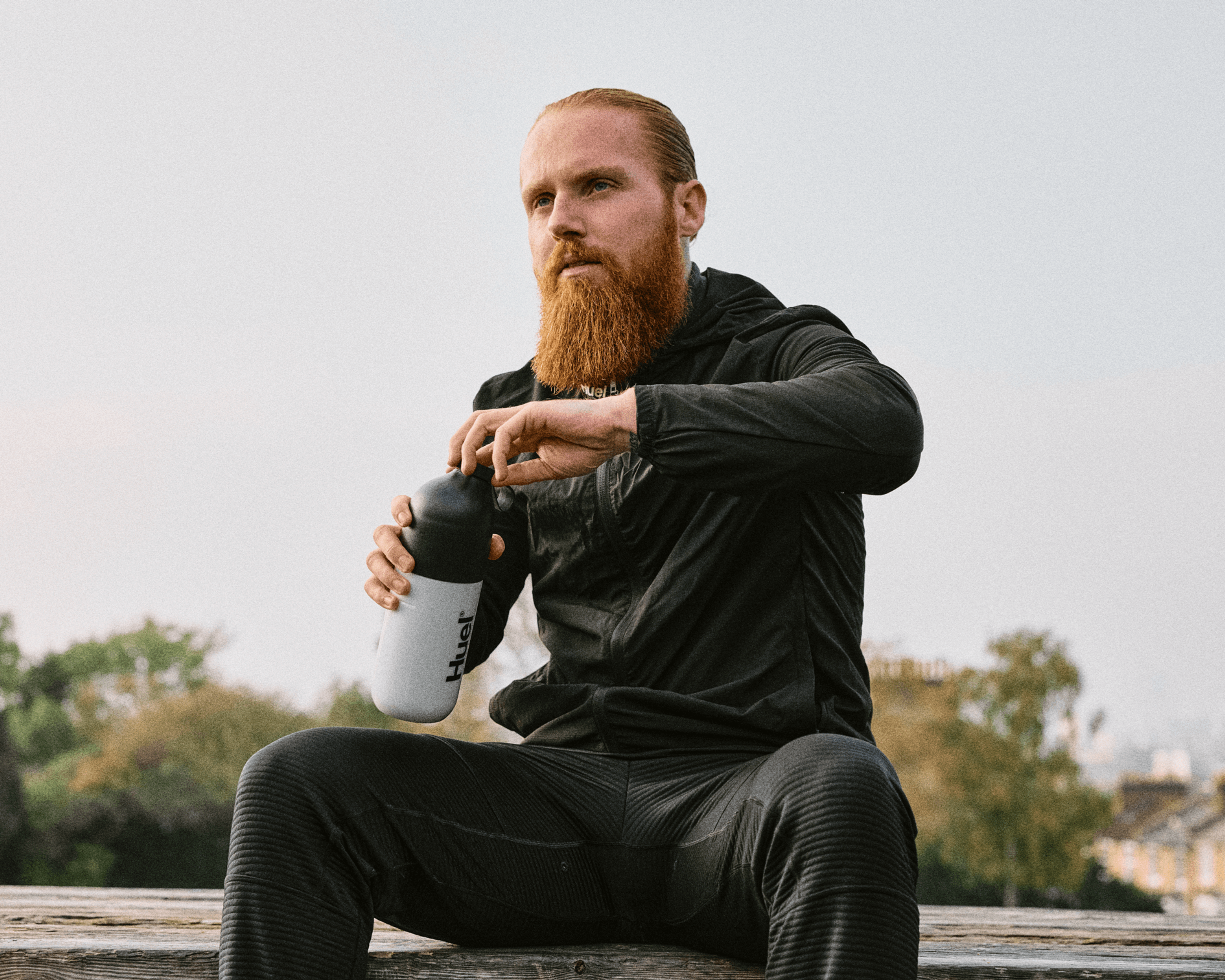 Russ Cook - The Hardest Geezer - sitting on a bench in a black tracksuit, opening a shaker of Huel