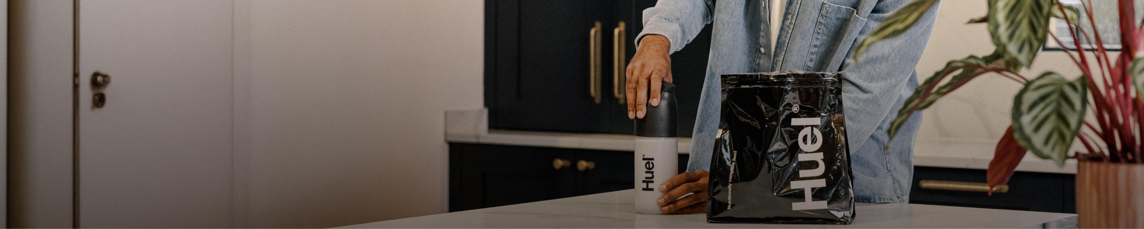 A man putting the lid on a black and white Huel shaker in a modern kitchen, next to a bag of black Huel powder