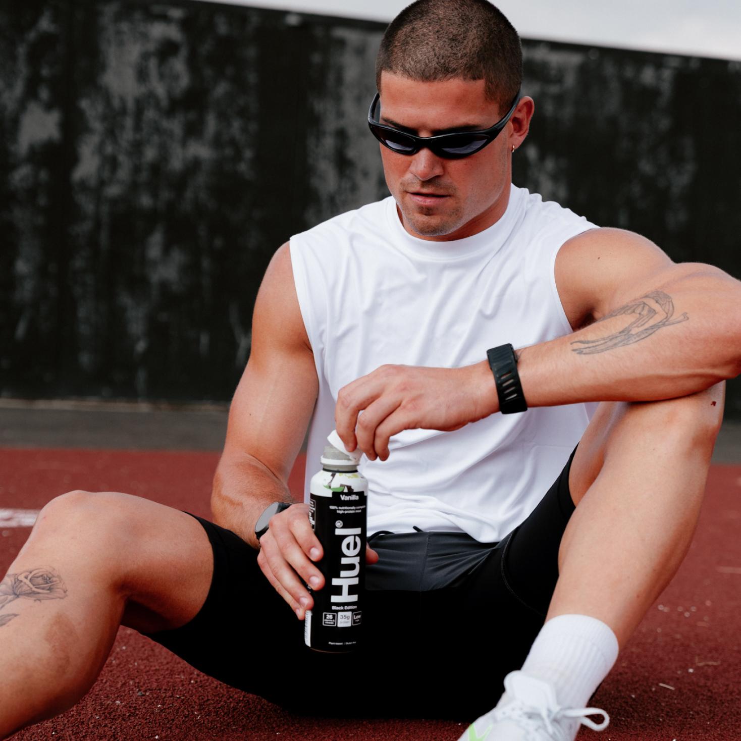 A man wearing a sleeveless white tank and black shorts is sitting on ground unscrewing the top off a Black Edition Ready-to-drink
