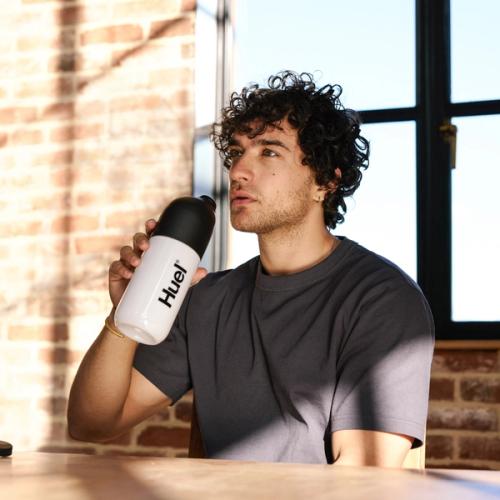 A man with curly hair is sitting indoors, sipping from a white Huel shaker with a black lid. He is seated at a wooden table in a sunlit room with large windows and exposed brick walls, enjoying a healthy meal replacement shake.