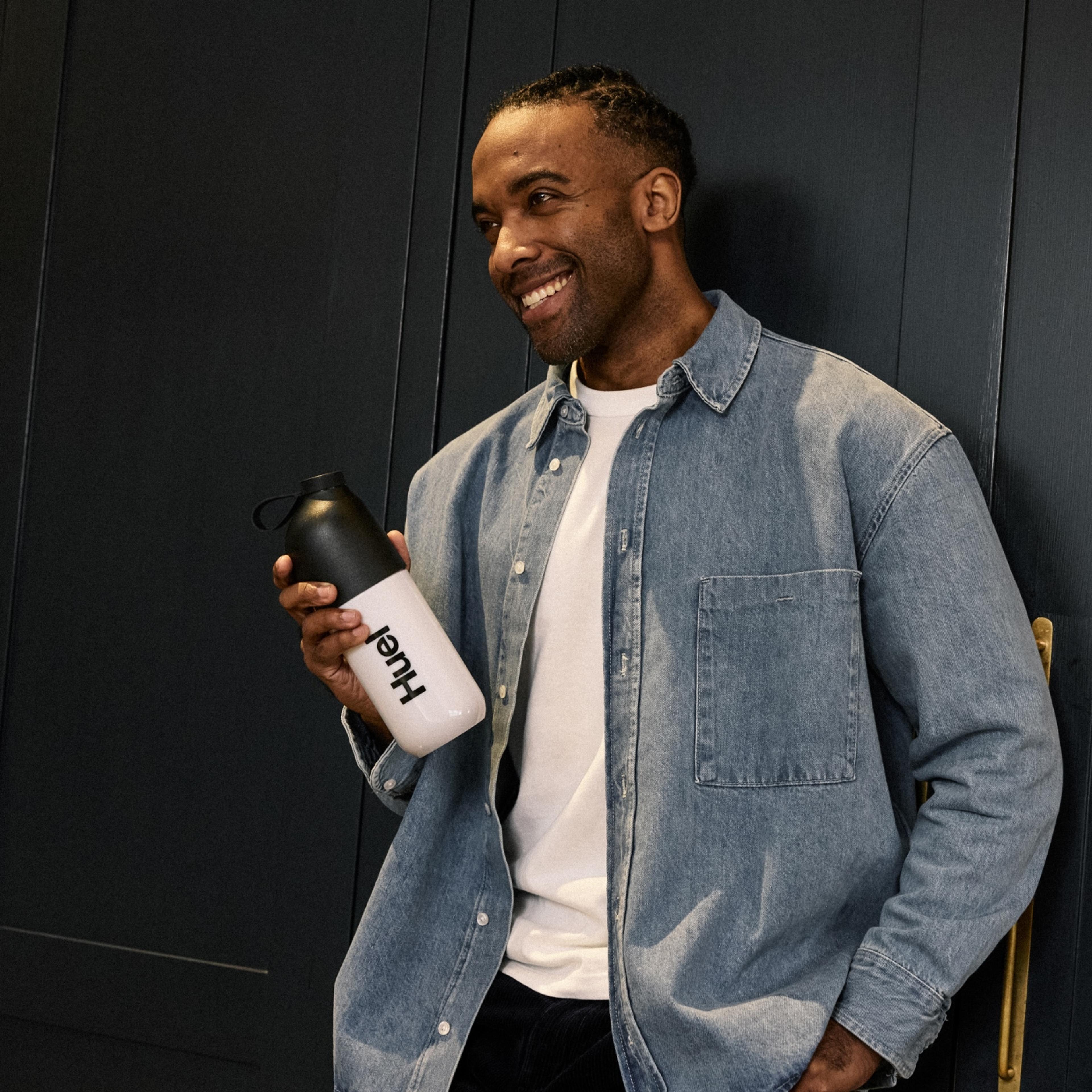 A smiling man in a denim jacket is holding a Huel Black Edition shaker bottle with a black lid. He stands in front of a dark wall, exuding a relaxed and confident vibe in a modern indoor setting.