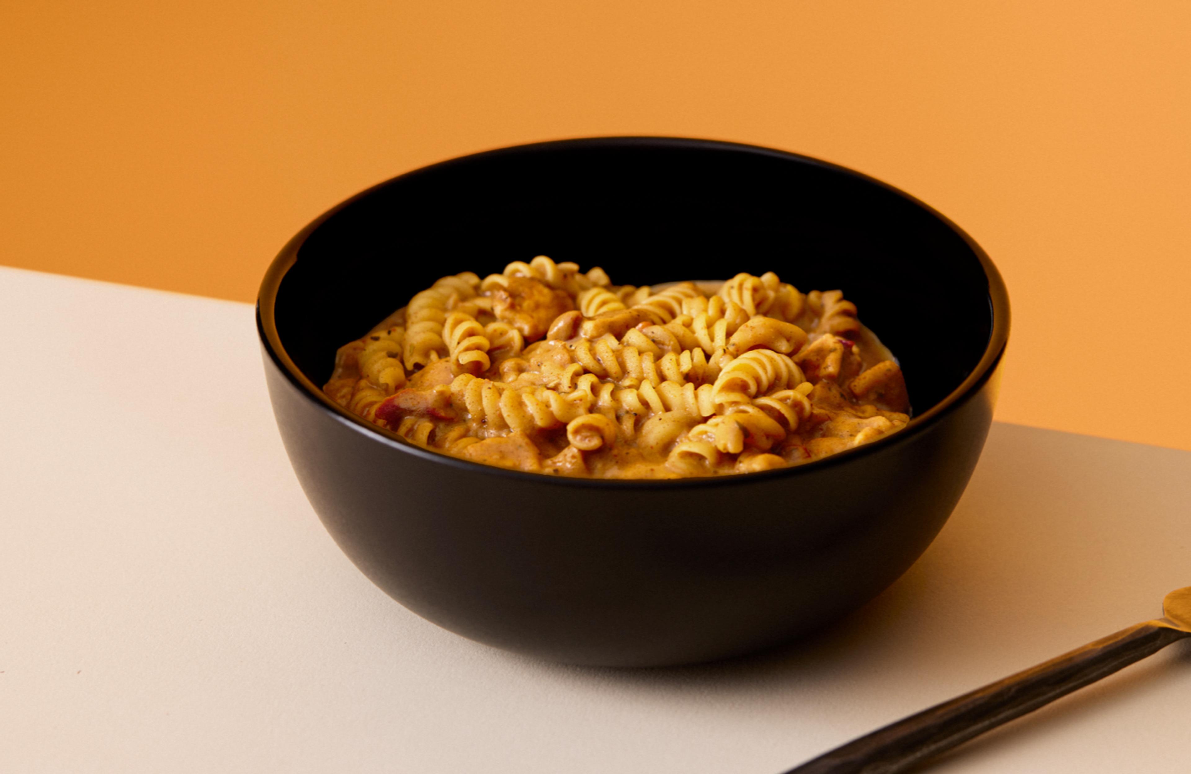 A bowl of Cajun Pasta Huel Hot & Savoury, on a table, set against a yellow background
