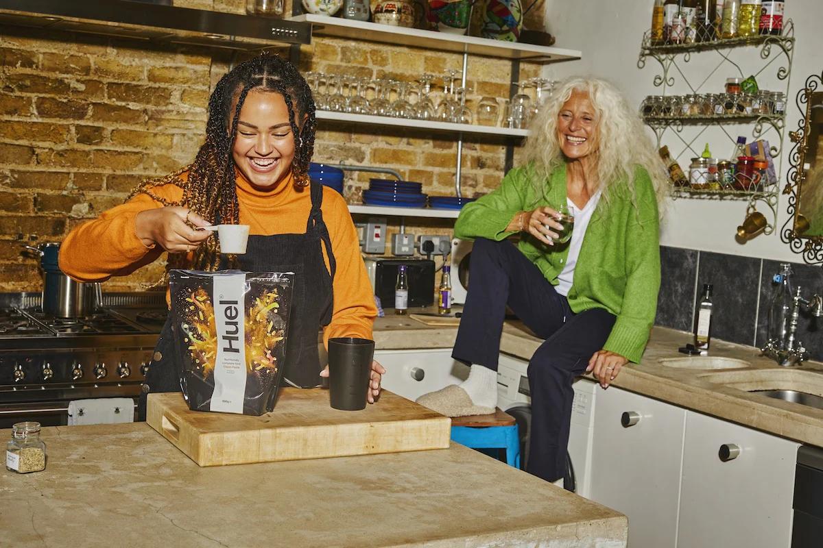 two women in a kitchen smiling one sat on the kitchen side and the other scooping huel hot and savoury