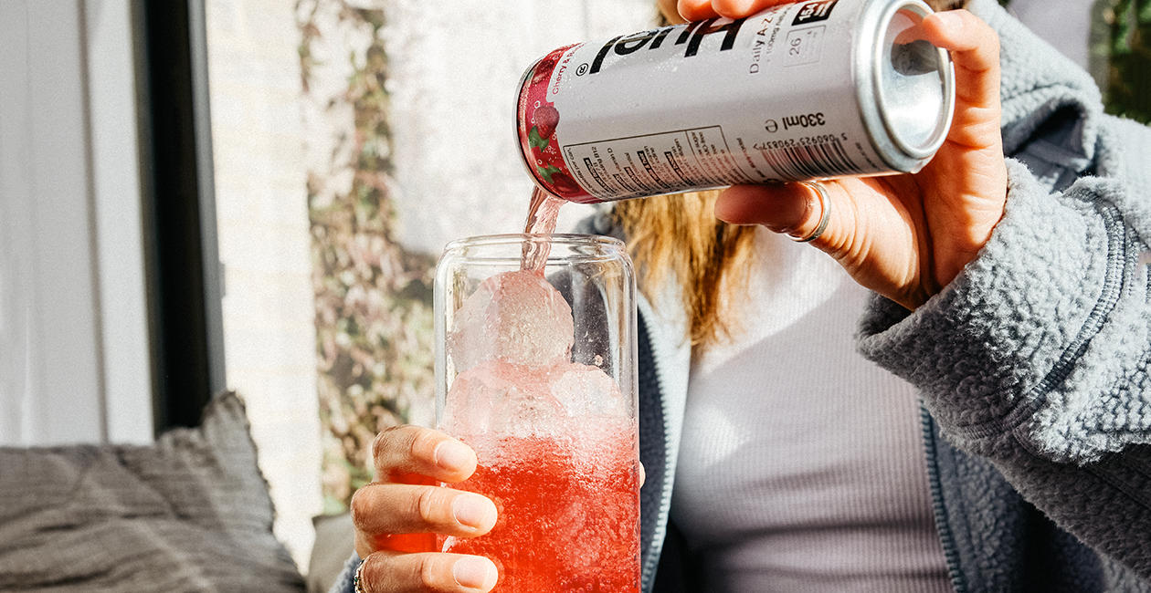 A lady pouring a can of Huel cherry & raspberry daily-a-z into a glass with ice