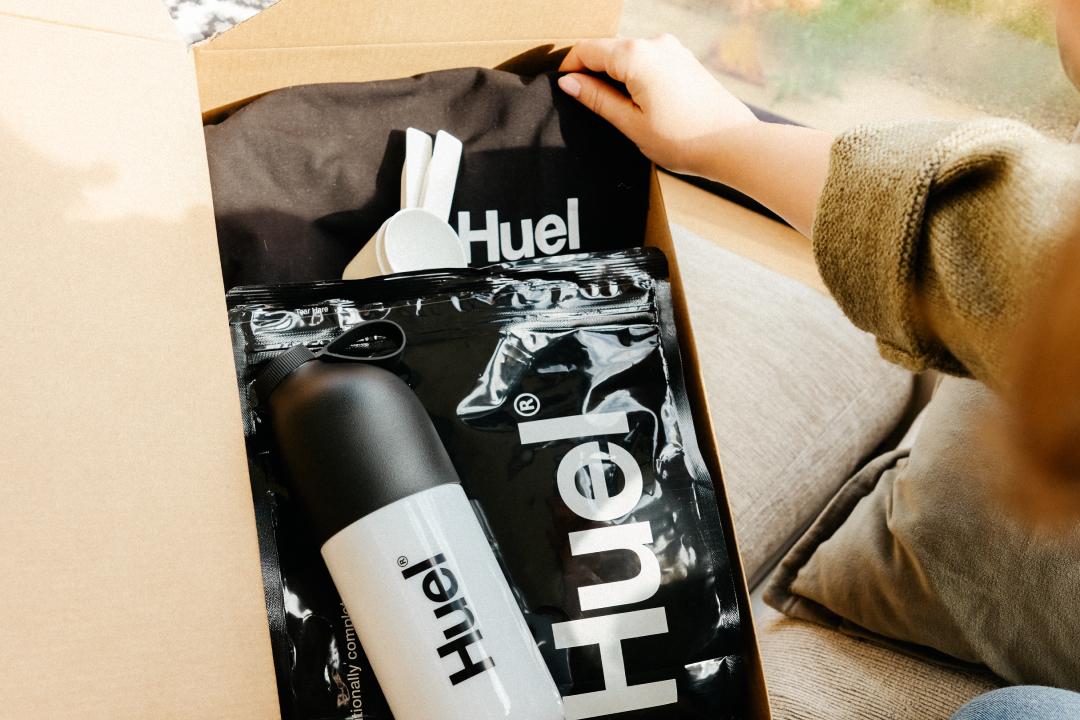 An open box contains Huel products, including a white and black shaker bottle, a large black pouch of Huel powder, a black Huel-branded T-shirt, and white scoops. A hand reaches into the box, which is placed near a window with natural light.