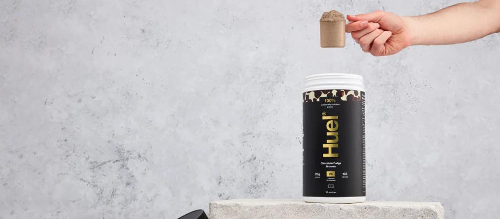 Huel Complete Protein tub with a scoop being held above it