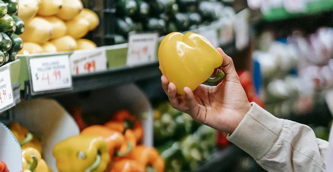 person in a supermarket holding a yellow bell pepper in the produce section