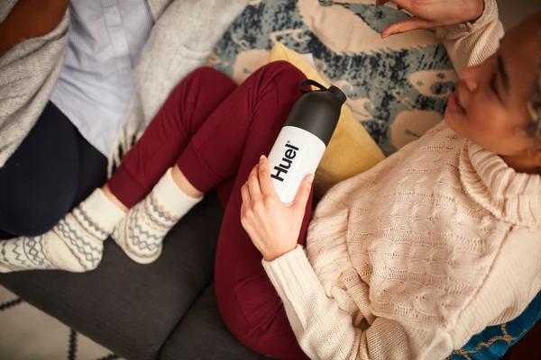 woman sat on a sofa holding a huel shaker against her leg