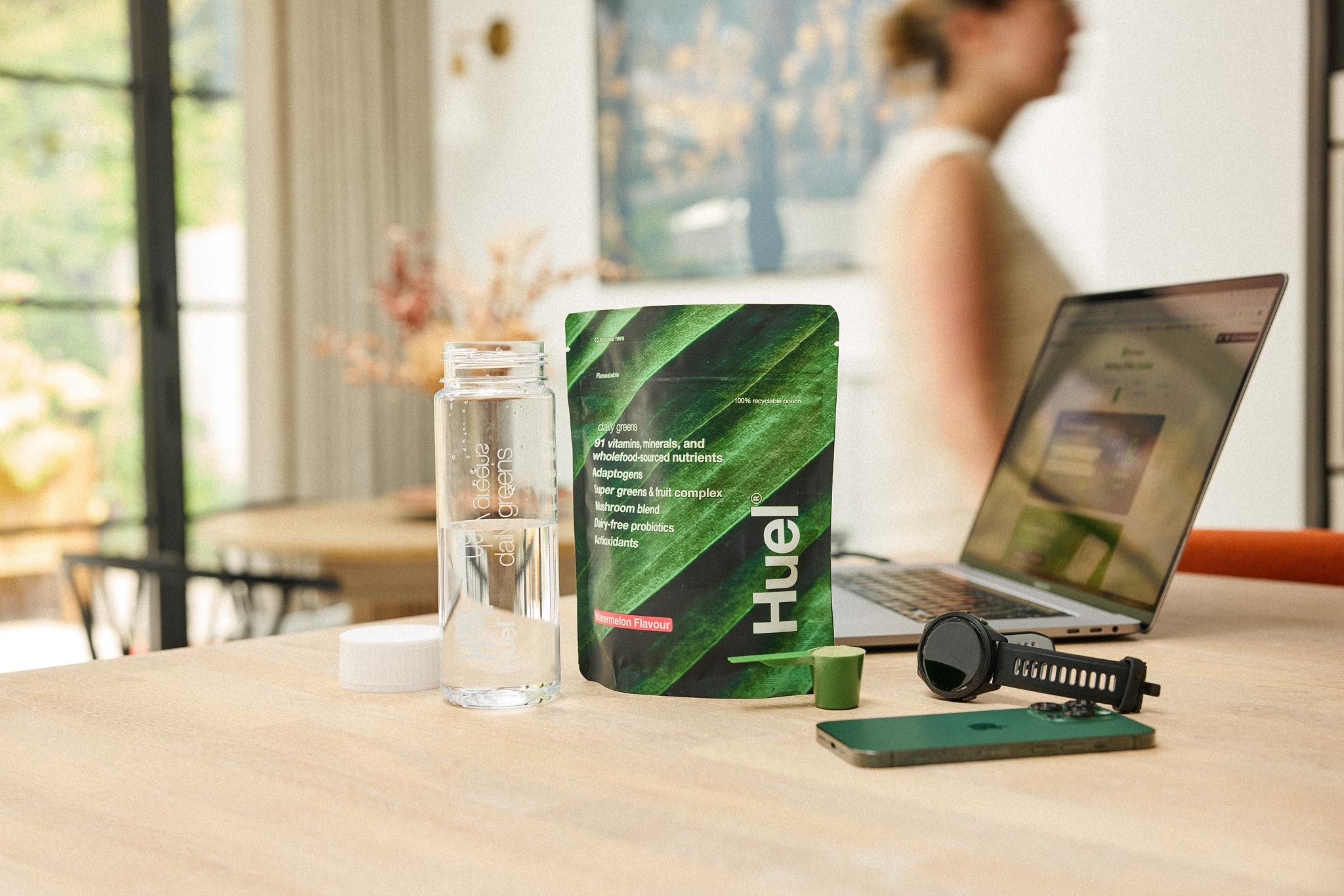 Huel Daily Greens pouch on a counter next to a smart watch phone and laptop