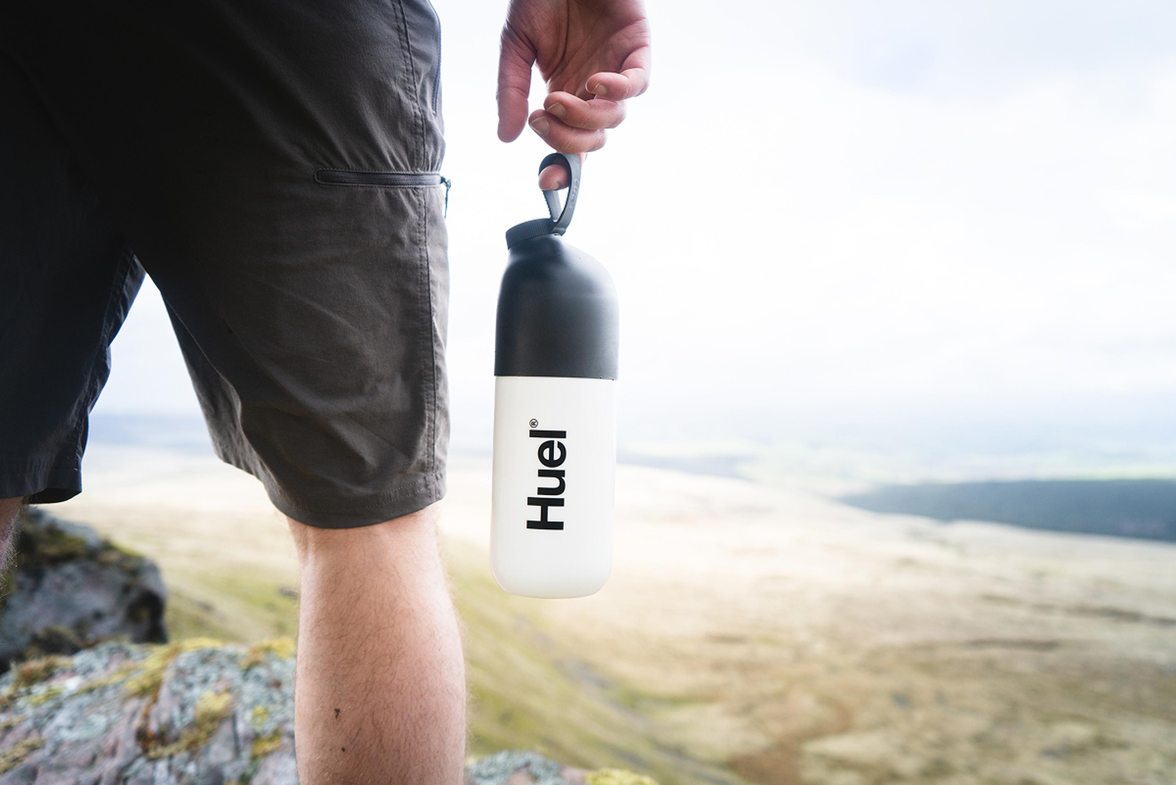 A Man holding a Huel shaker by the loop while outside in the mountains