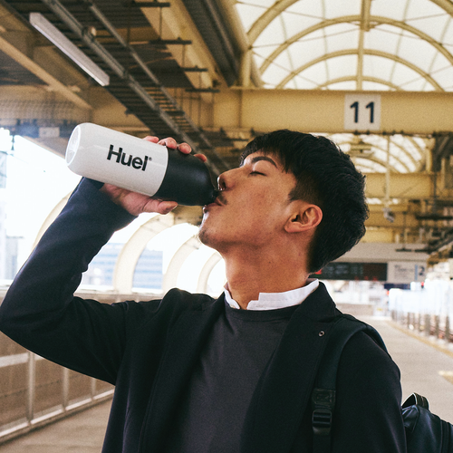 青空を背景に、白いボトルでHuel Ready-to-drinkを飲む若い女性。