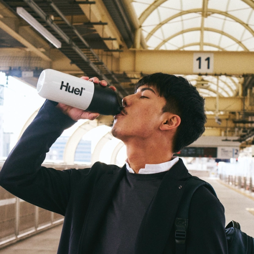 青空を背景に、白いボトルでHuel Ready-to-drinkを飲む若い女性。